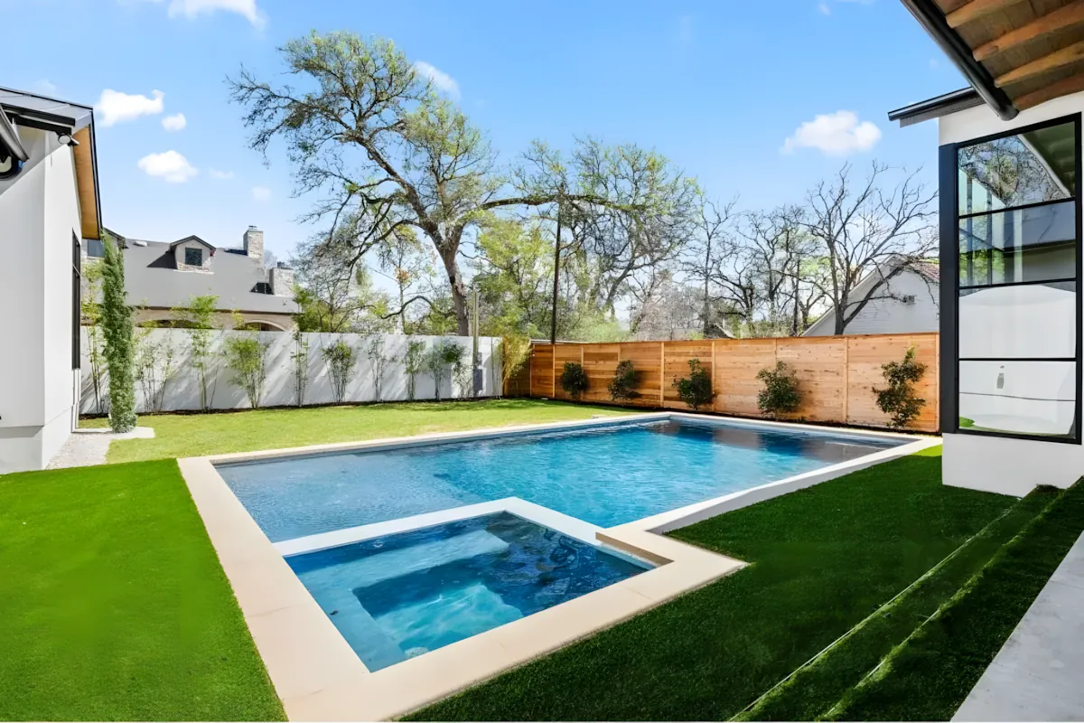 Pool with a square spa and turf lawn behind a stone home