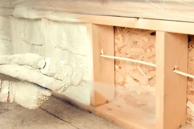 Gloved hand spraying off-white foam insulation into open wooden wall stud framing
