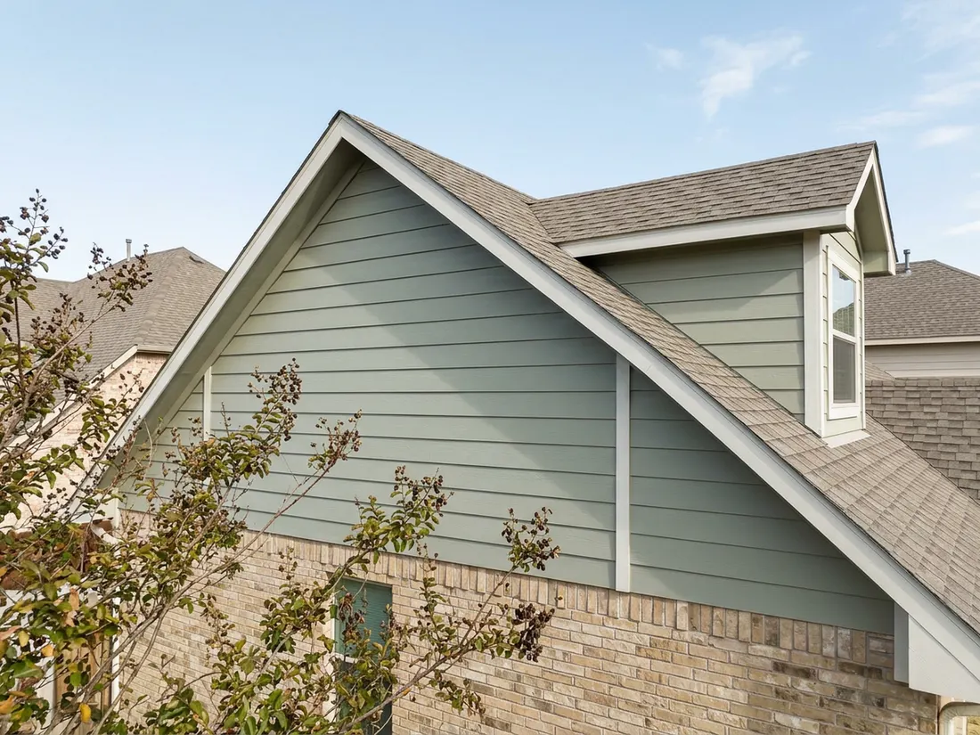 Sage fiber cement siding on a gable and dormer above tan brick on a Texas house