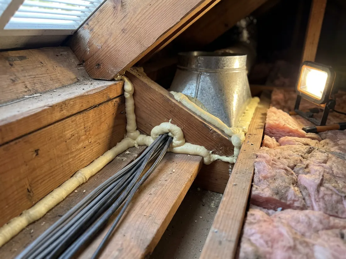 Close-up of spray foam sealing gaps around wiring and a duct boot at attic eave