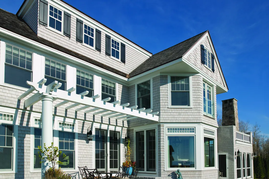 Gray cedar shake house featuring white-trimmed windows, black shutters, and an attached white pergola patio