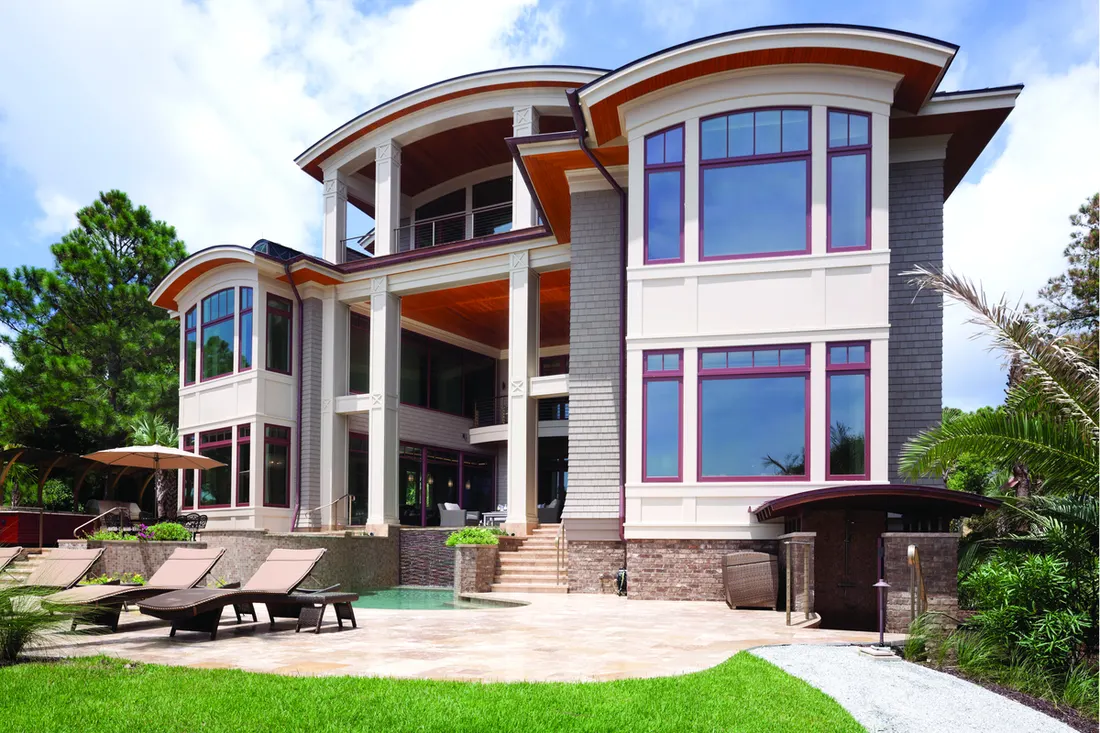 Large multi-story home with expansive red-trimmed windows, curved rooflines, patio loungers, and pool