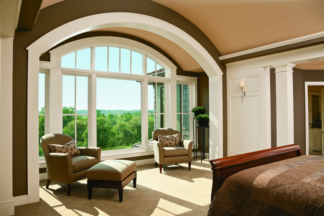 Large white arched window in a brown bedroom framing views of green outdoor trees