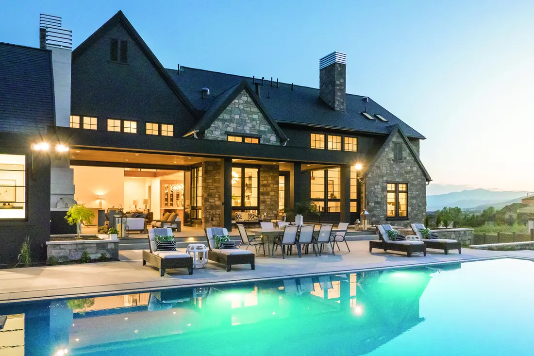 Illuminated swimming pool and patio beside a modern dark home with stone accents at dusk
