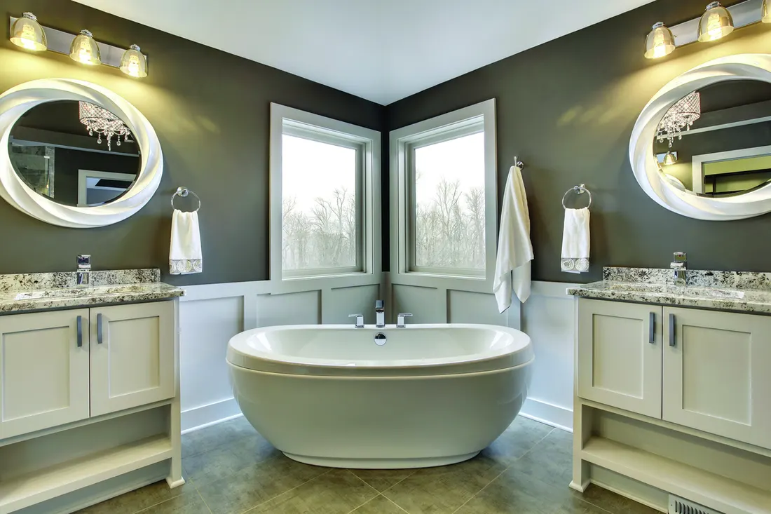 Two trimmed corner windows behind a white freestanding tub in a gray and white bathroom