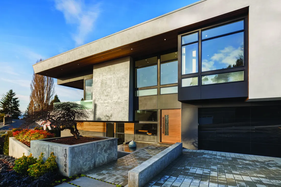 Modern home exterior featuring large black-framed picture windows, concrete walls, and a wooden front door