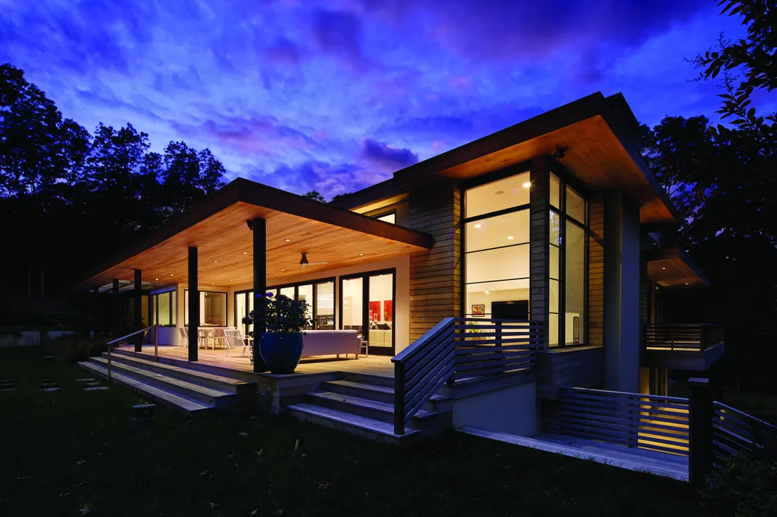 Modern home exterior at dusk featuring large floor-to-ceiling glass windows and illuminated patio doors