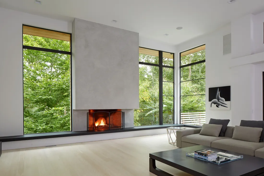 Large floor-to-ceiling black frame windows beside a modern fireplace overlooking green outdoor trees