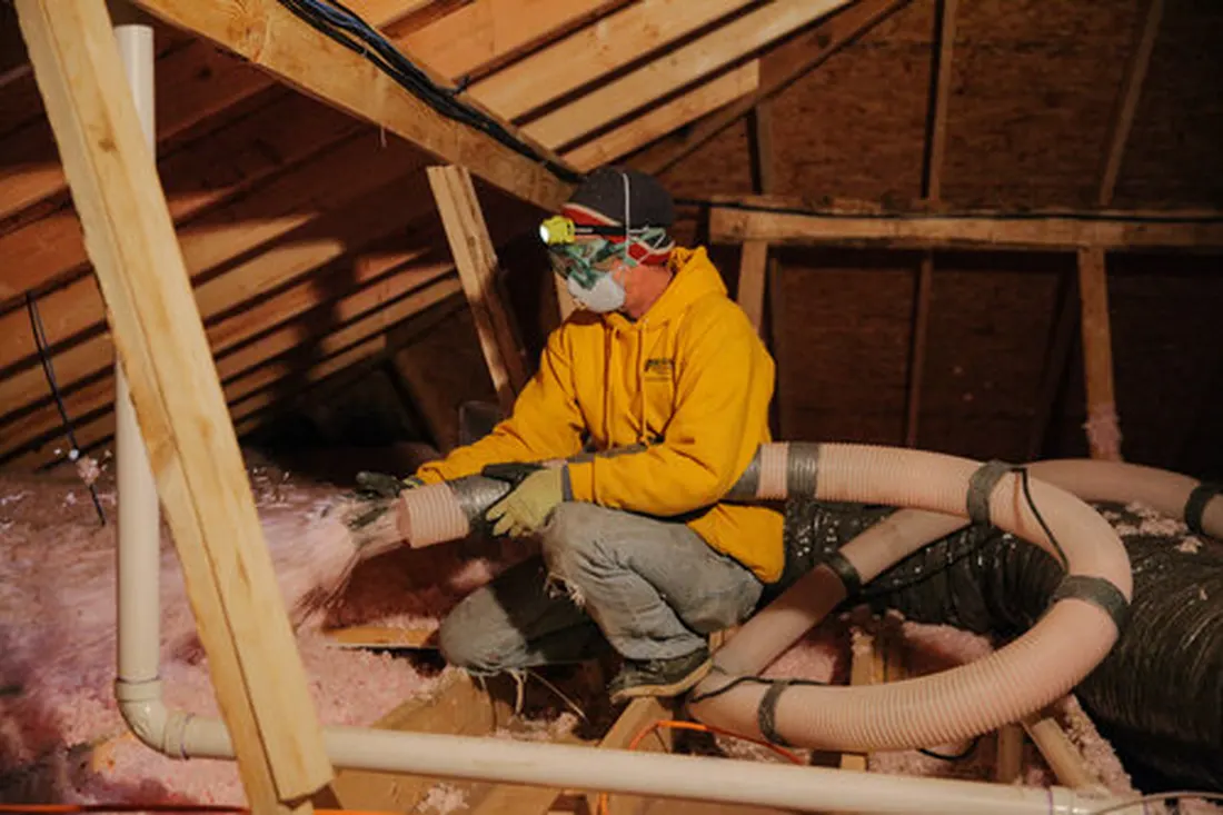 Worker in yellow hoodie spraying pink insulation through a flexible hose in an attic