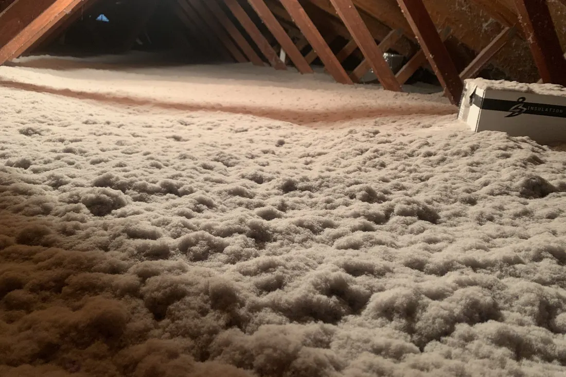 Thick layer of fluffy white blown-in insulation covering an attic floor beneath wooden roof trusses
