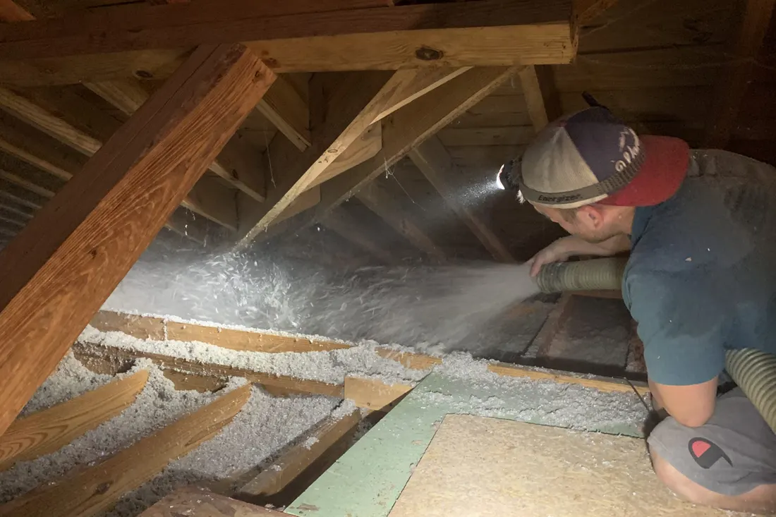 Worker spraying white blow-in insulation between wooden joists and beams inside a dark attic