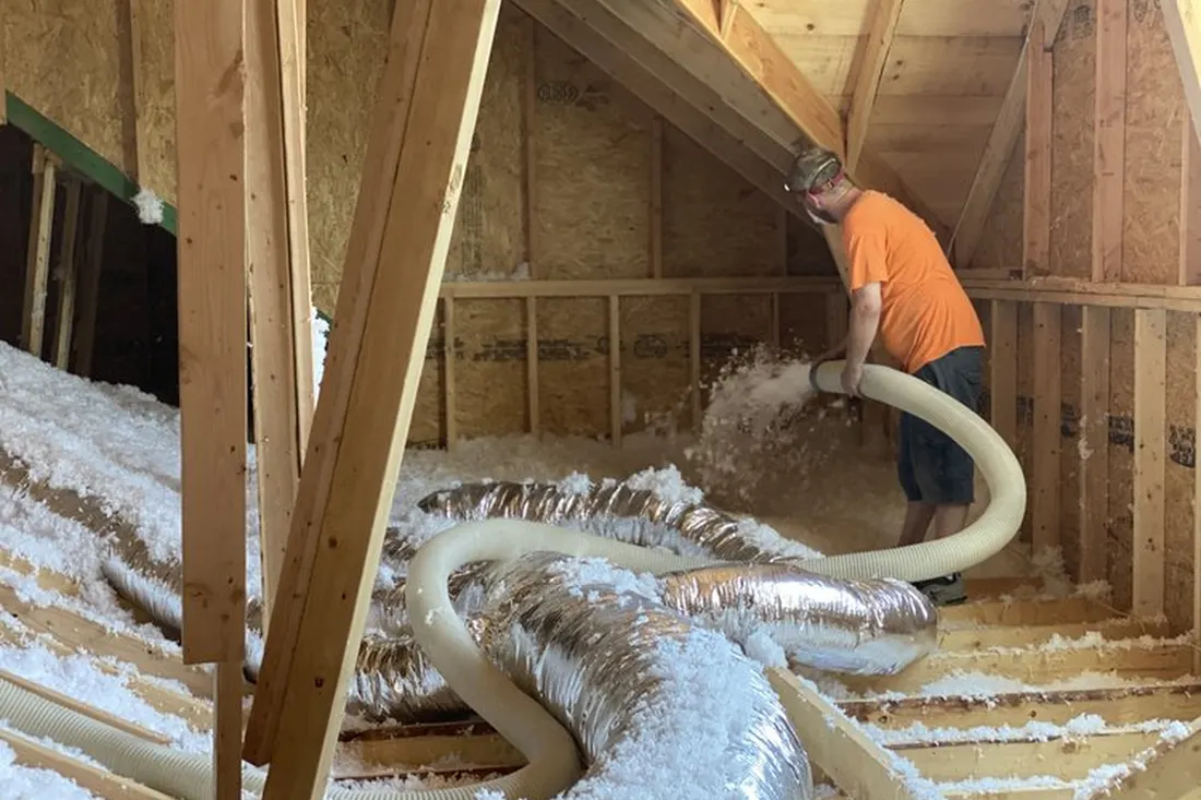 Worker spraying white blow-in insulation from a hose across wooden attic rafters and silver ductwork