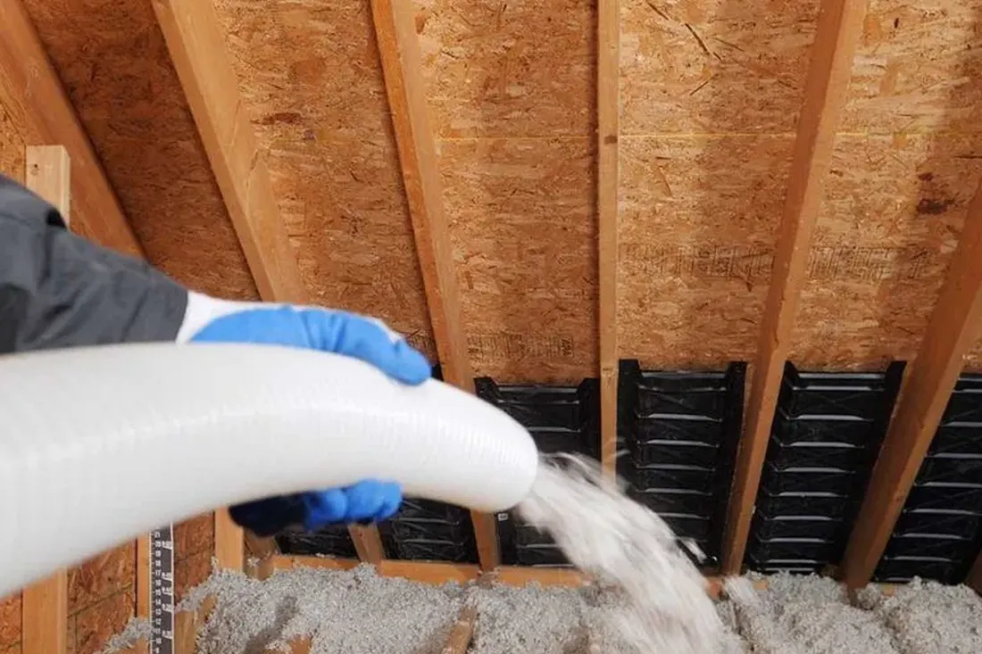 Gloved hand spraying blown-in insulation from a white hose onto an attic floor