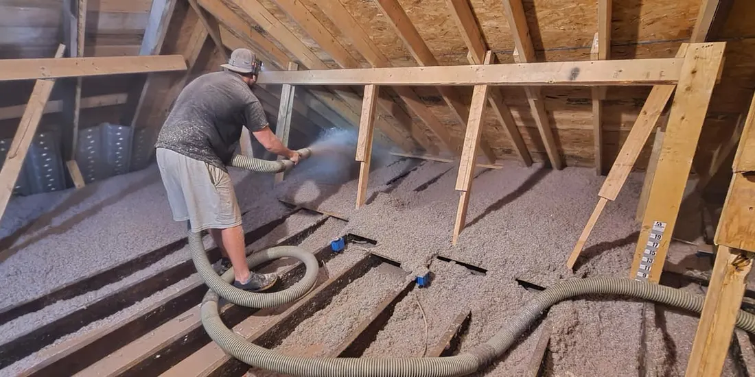 Worker spraying gray blow-in insulation between wooden joists in an attic space under rafters