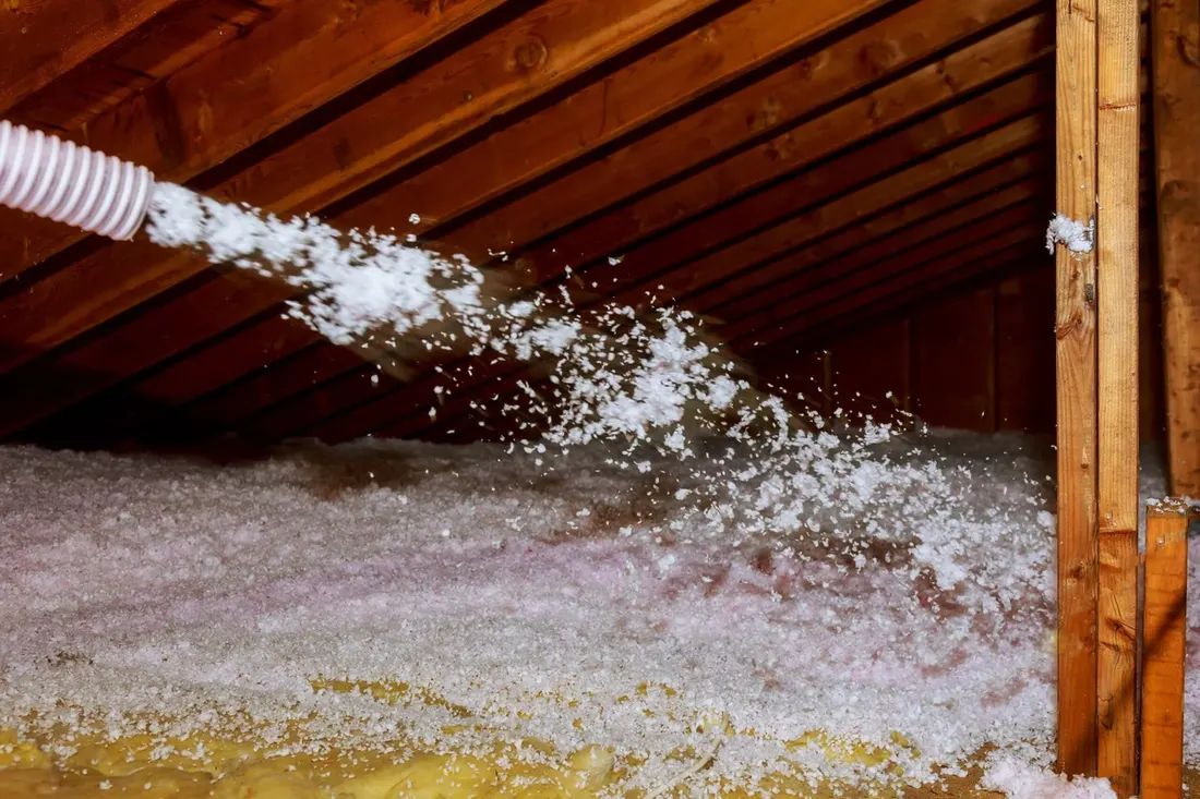 White flexible hose spraying loose white insulation across the floor of a wooden attic
