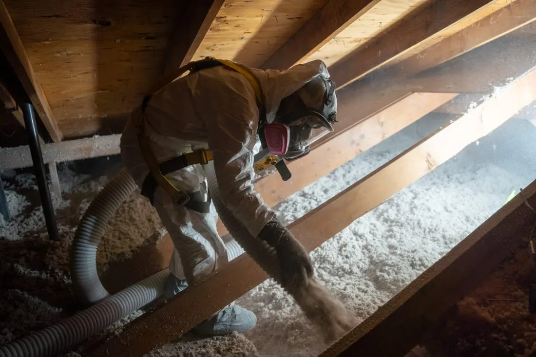 Worker in protective gear spraying white blown-in insulation between wooden attic rafters using a hose