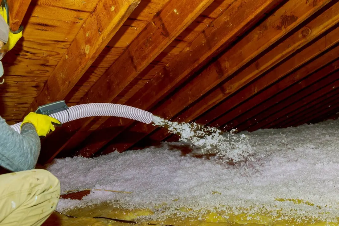 Worker spraying white blown-in insulation onto an attic floor beneath exposed wooden rafters