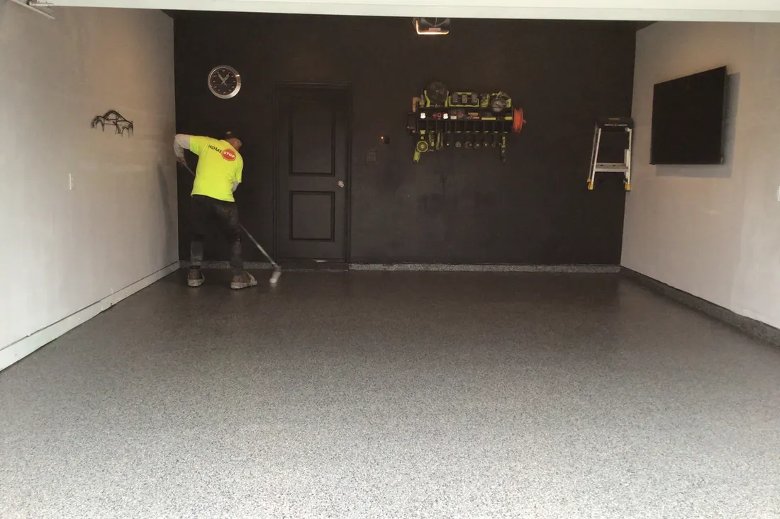 Worker applying glossy gray speckled concrete coating to a residential garage floor with dark walls