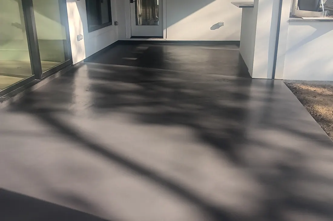 Smooth dark gray coated concrete patio with tree shadows outside a modern white house