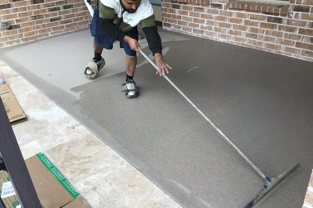 Worker spreading a smooth gray concrete coating across an outdoor patio with brick walls