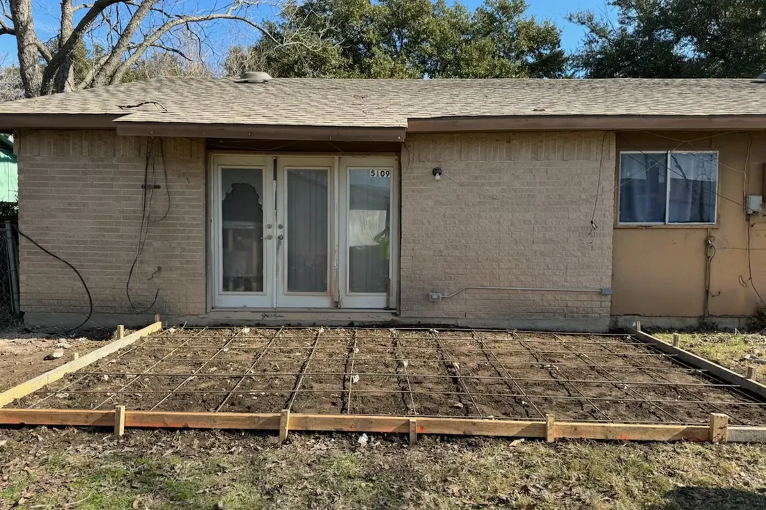 Wooden framing and rebar grid on dirt ground behind a tan brick house