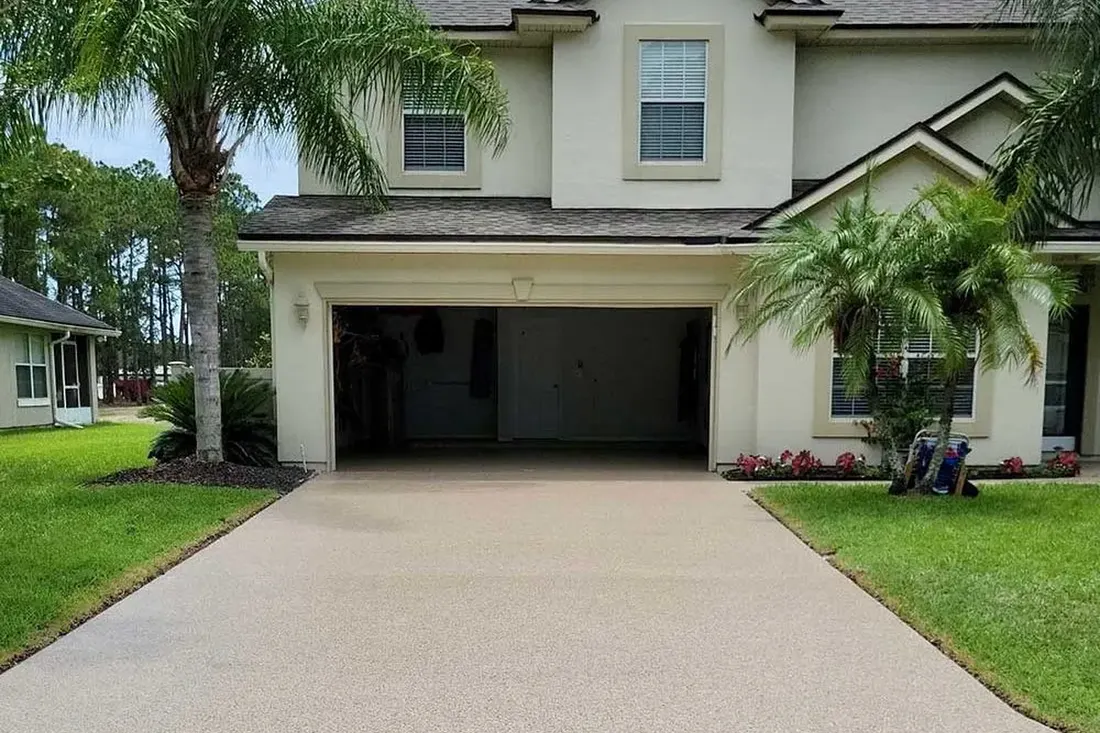 Beige coated concrete driveway leading to an open garage on a two-story home with palm trees