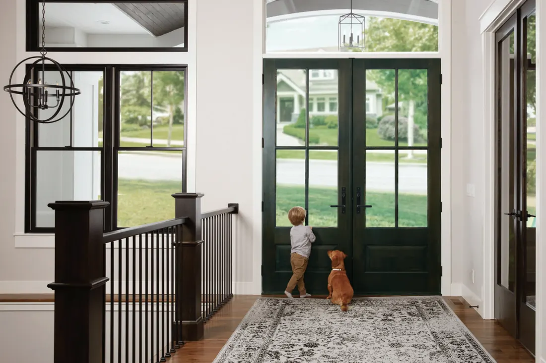 Double glass-paneled dark green entry doors with transom window and black handles in a bright foyer