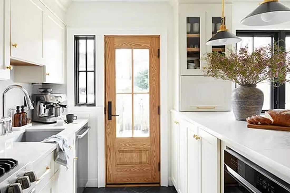 Natural wood entry door with glass panes centered between white kitchen cabinets and countertops