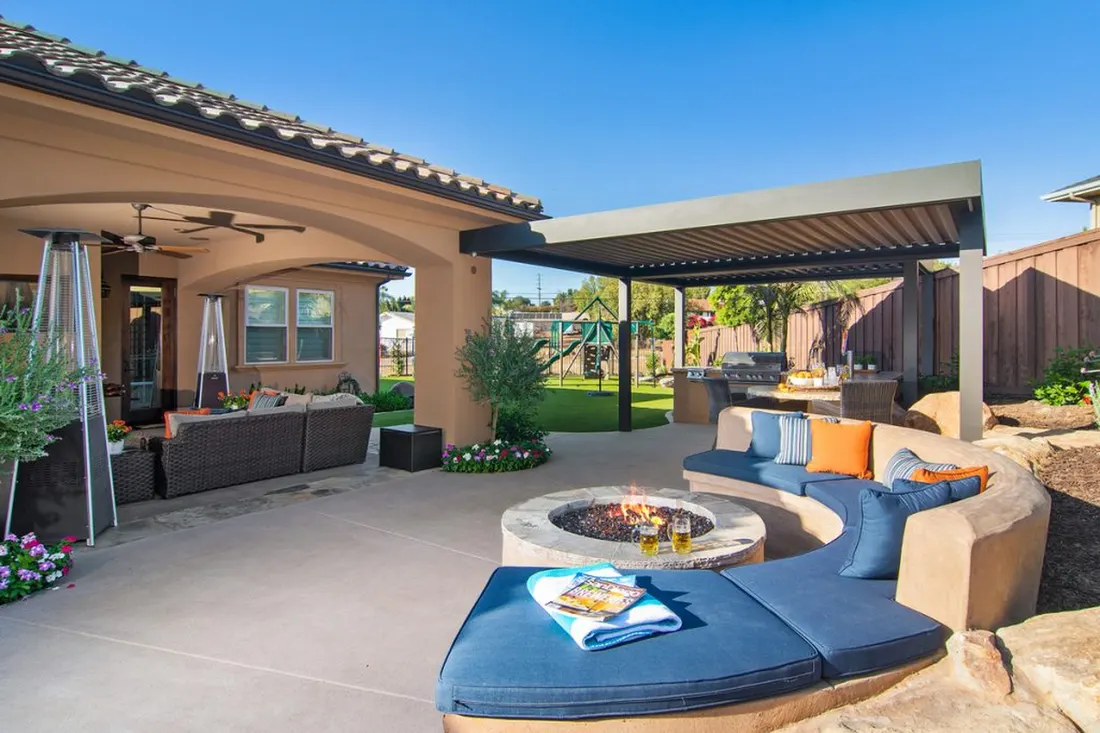 Patio with round fire pit, curved blue sofa, covered pergola kitchen, and sunny backyard lawn