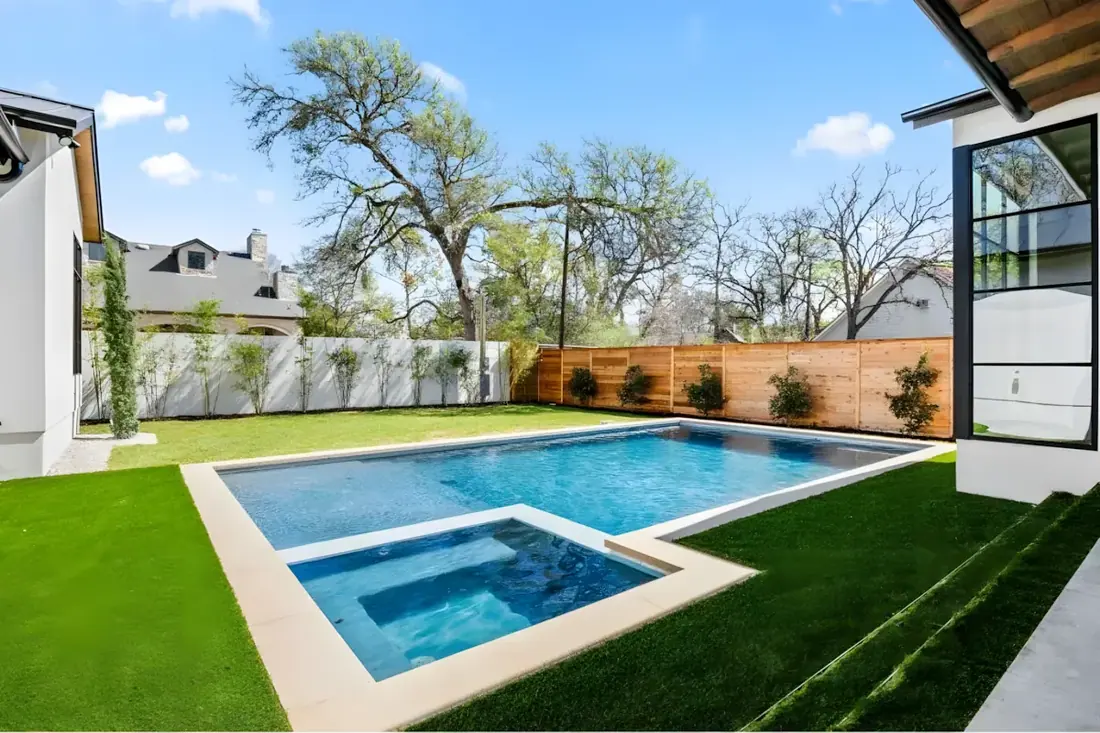 Pool with a square spa and turf lawn behind a stone home