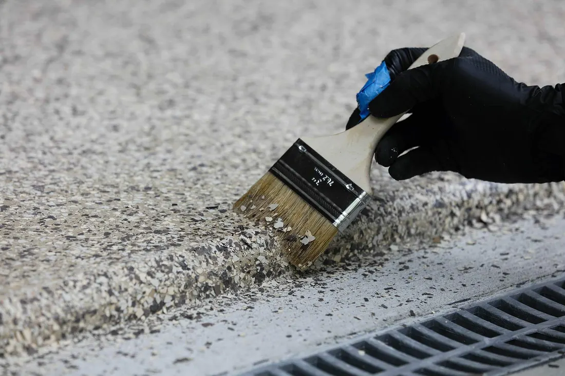 Gloved hand using a paintbrush to apply speckled flake coating along a concrete floor edge