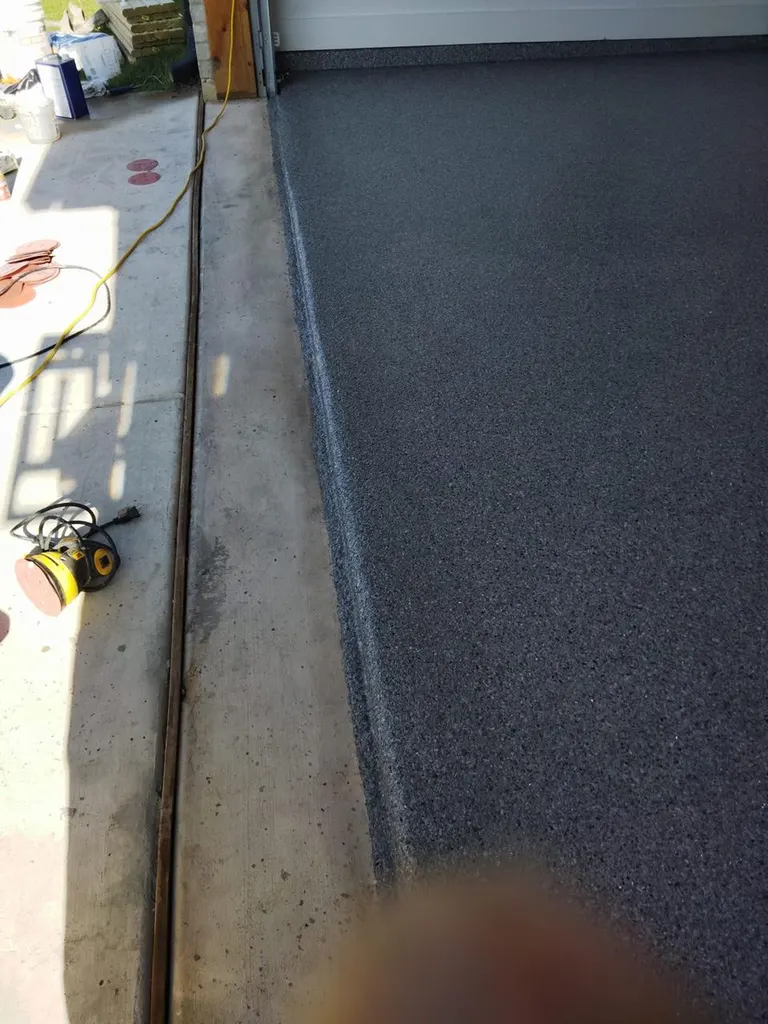 Dark gray textured epoxy coating applied to a garage floor beside unfinished concrete driveway threshold