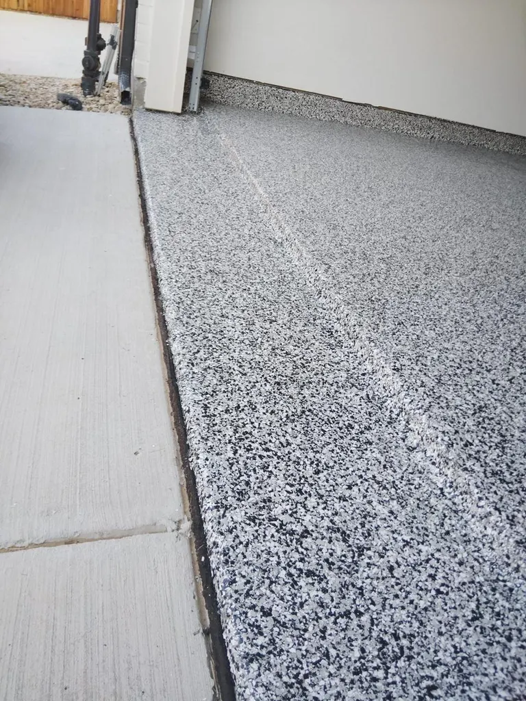 Garage entrance featuring speckled black, gray, and white coated floor adjacent to plain concrete driveway