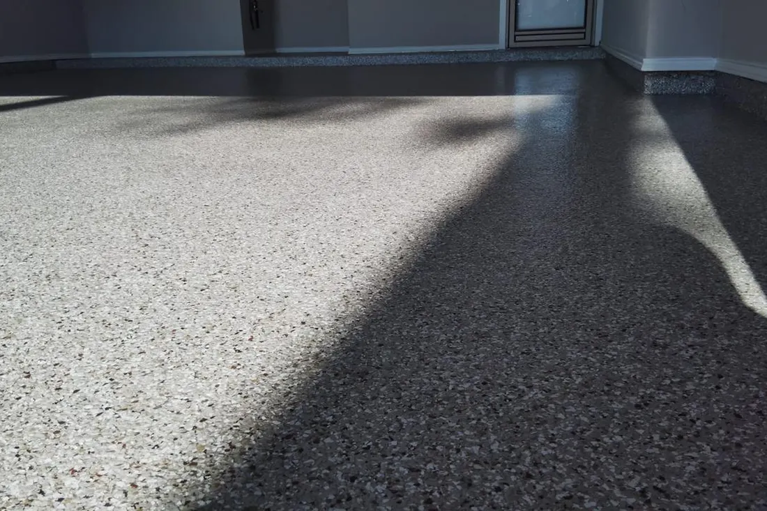 Glossy gray speckled epoxy coating applied across an indoor garage floor with sunlight and shadows