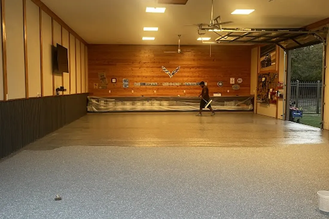 Worker applying a glossy clear coat over a speckled gray floor in a spacious garage