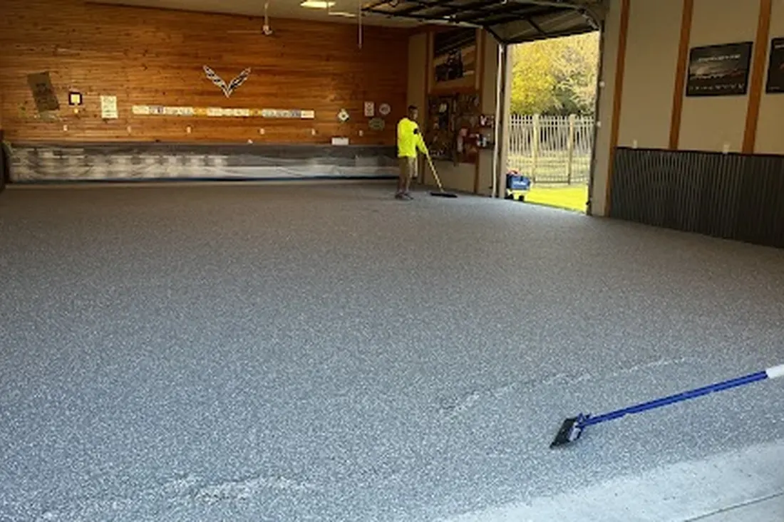 Worker applying gray textured epoxy coating inside a spacious garage with wood-paneled walls