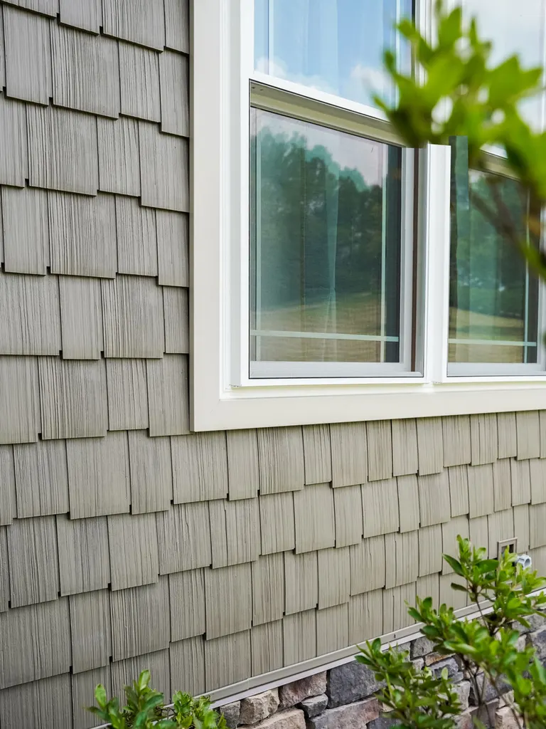 Gray textured shake siding around a white-trimmed window above a stone foundation base