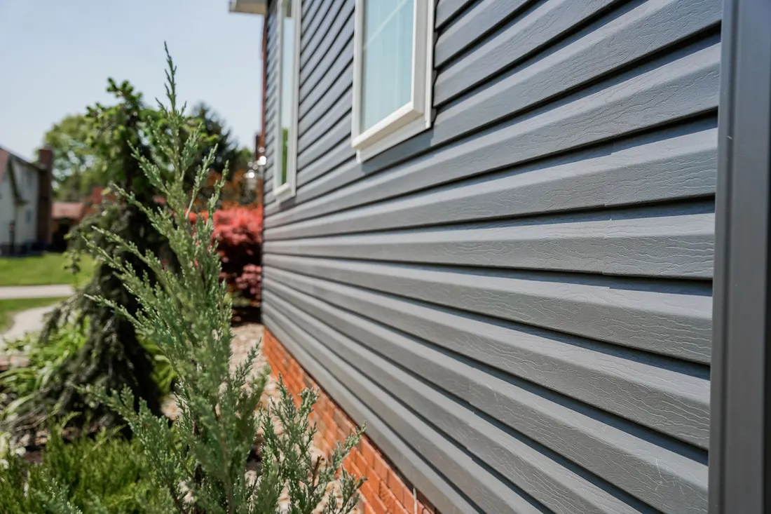 Gray horizontal siding installed on a house exterior above a red brick foundation beside green shrubs