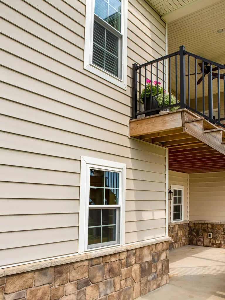 Tan horizontal siding and stone veneer foundation with white-framed windows beside a second-story deck