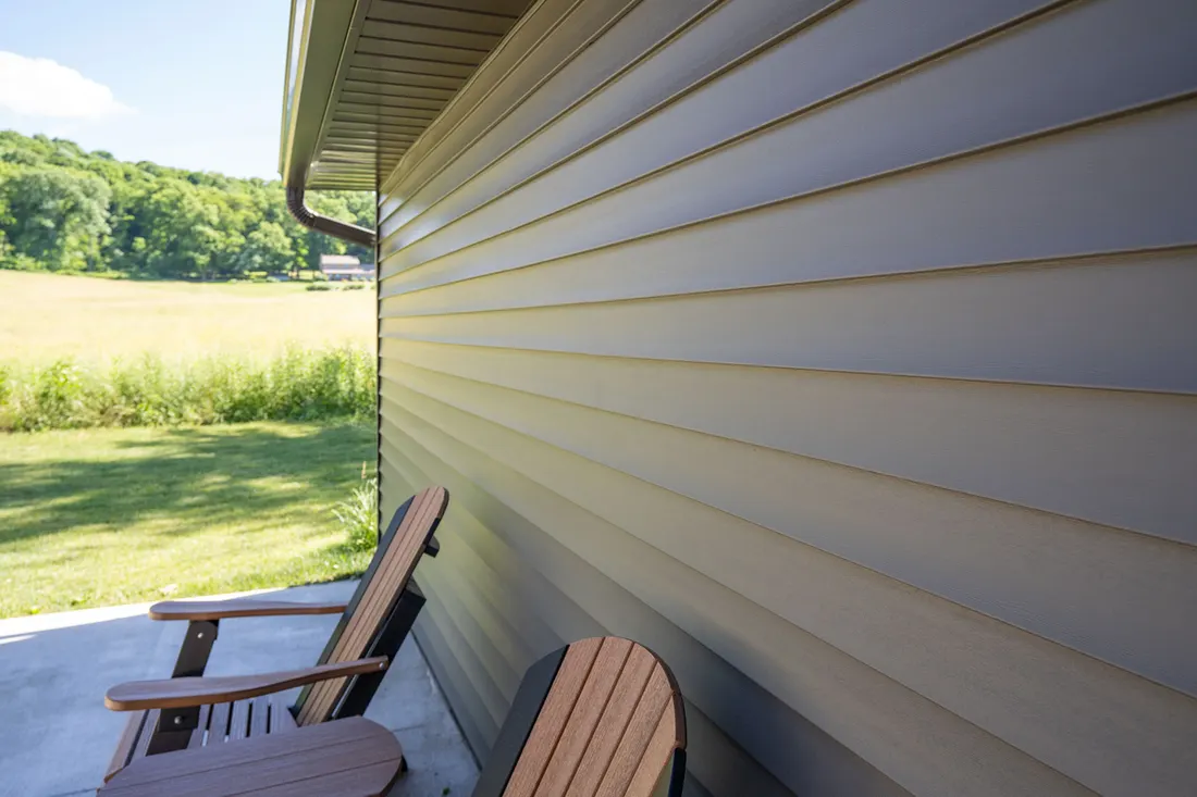 Light gray vinyl siding along a porch overlooking a sunlit grassy field and tree-lined landscape