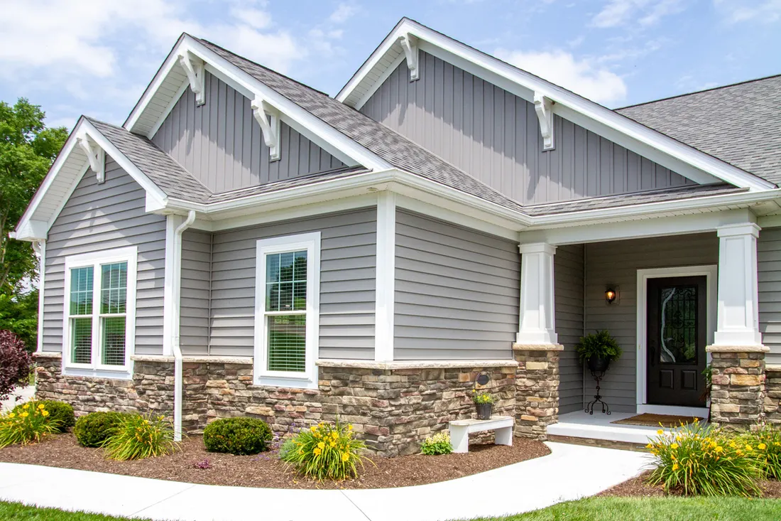 Gray vinyl siding home with white trim, stone accents, gabled roof, landscaping, and covered entryway