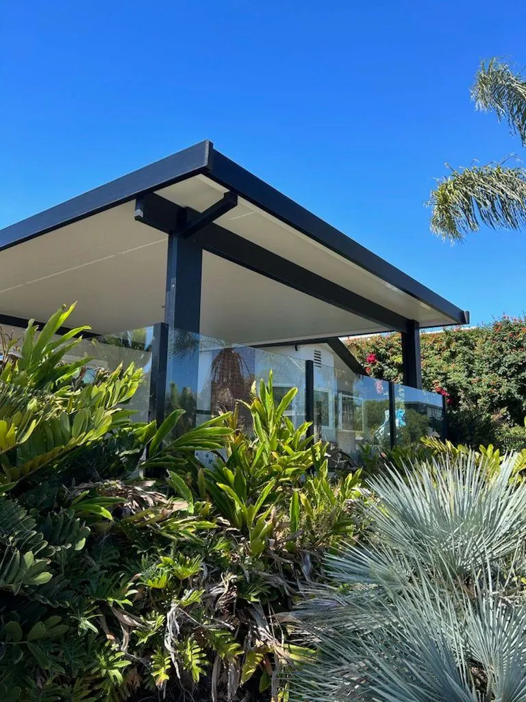 Modern black patio cover with glass railing surrounded by tropical plants under clear blue sky