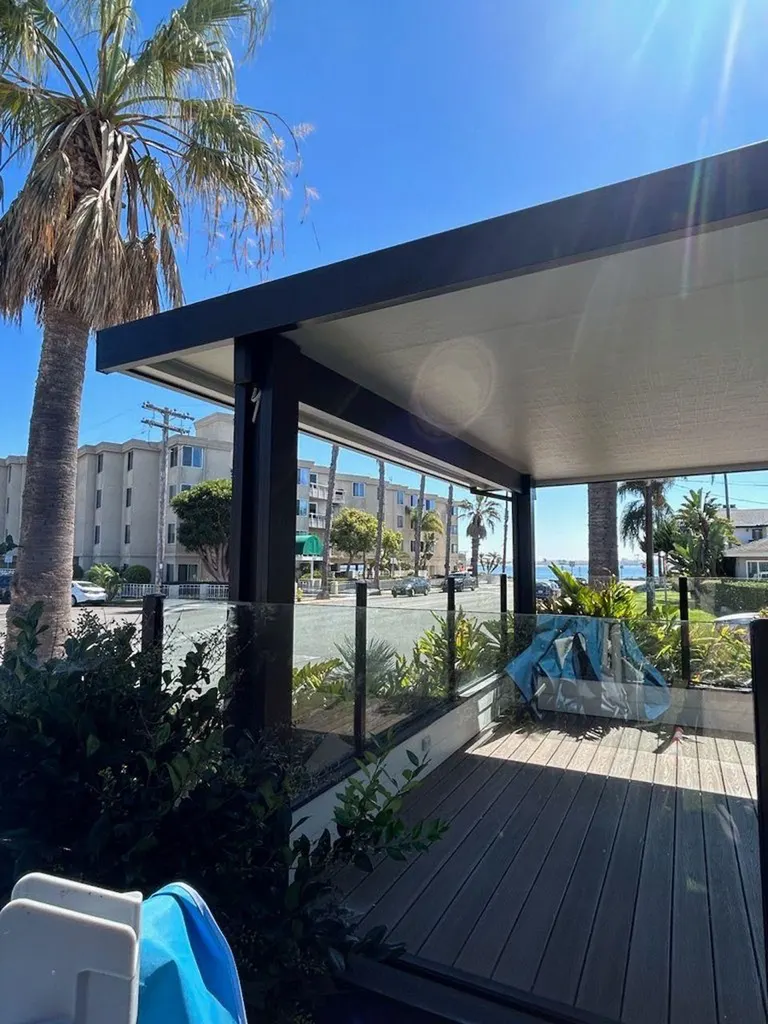 Modern dark-framed patio cover with white ceiling, glass railing, and wood decking beside palm trees