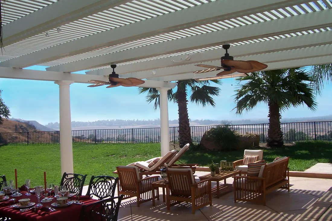 White slatted patio cover with ceiling fans, wood furniture, palm trees, lawn, and mountain views