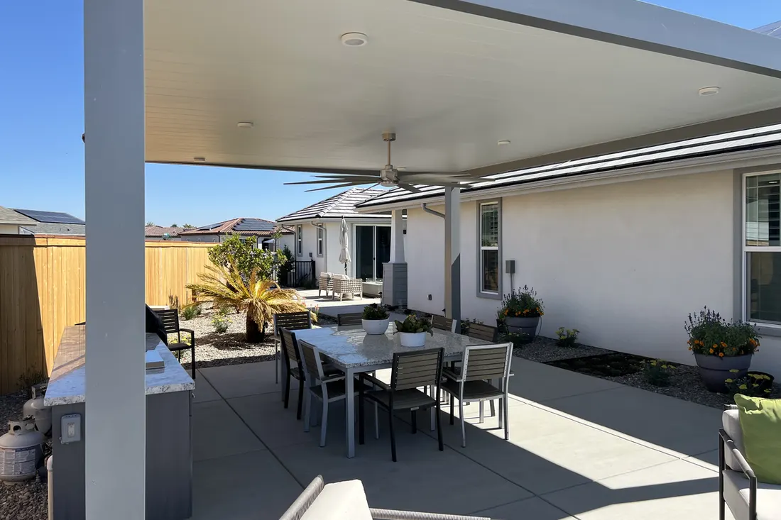 Covered patio with white ceiling, gray posts, ceiling fan, dining table, and concrete flooring beside a house