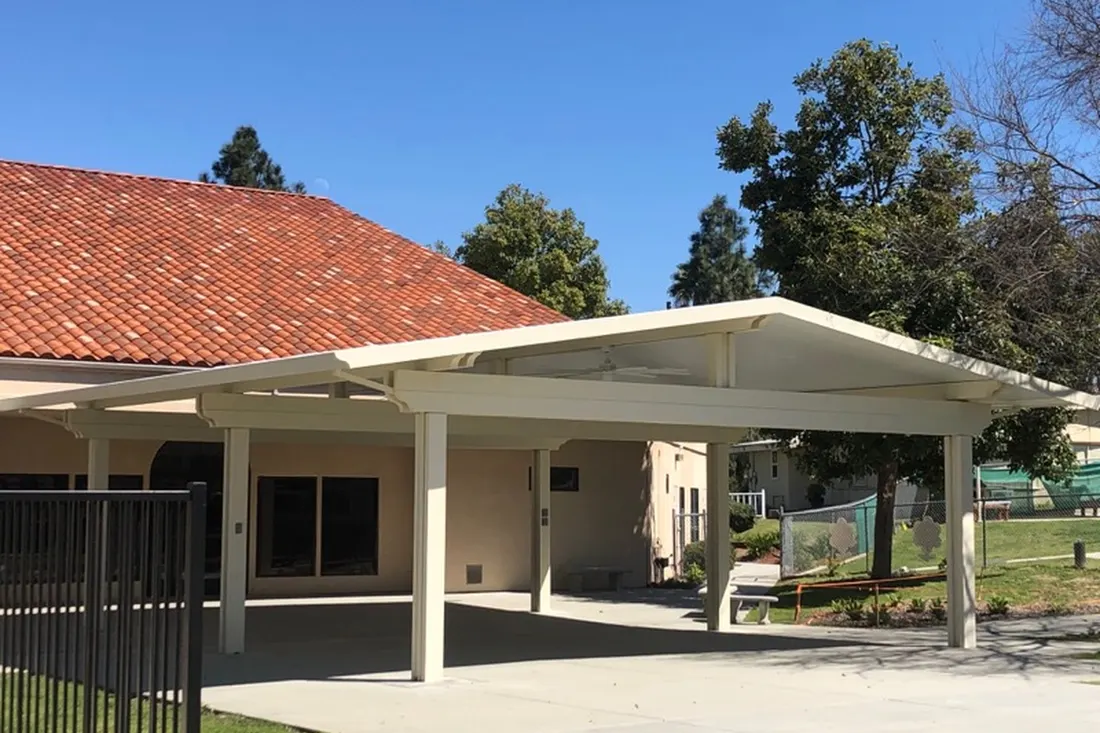 Cream-colored patio cover with sloped roof beside a beige home, concrete flooring, trees, and fenced yard