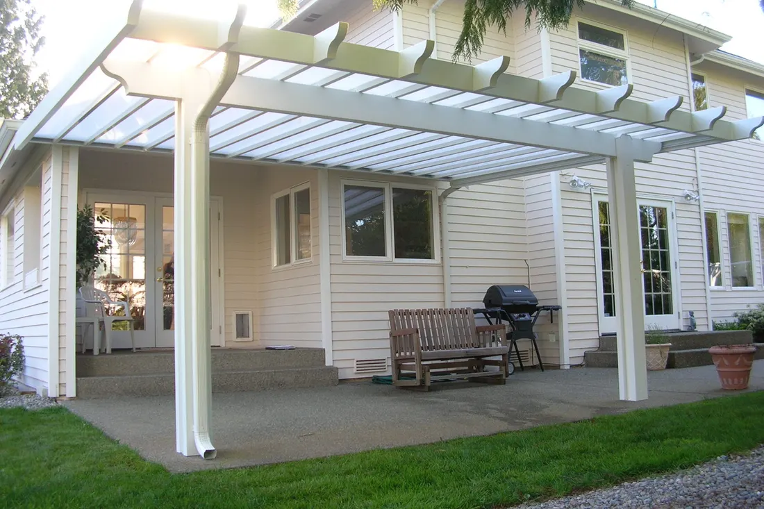 White patio cover with translucent roof panels, supported by posts beside a beige-sided house and lawn