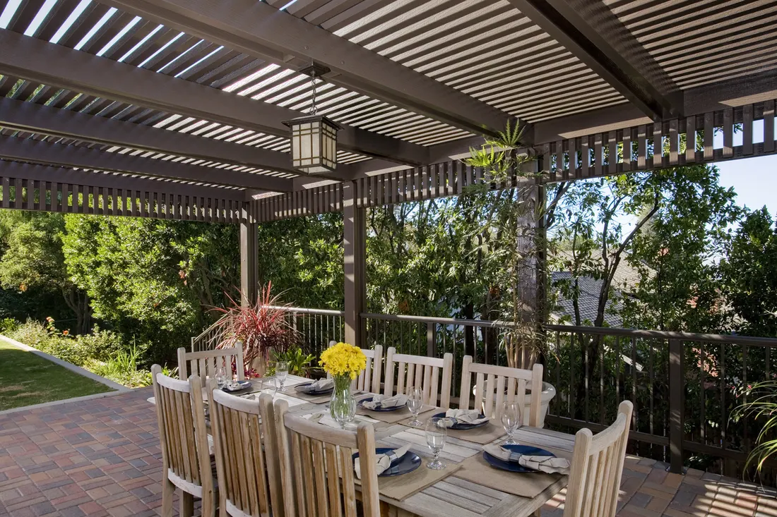 Dark-stained wood patio cover with slatted roof over an outdoor dining area, surrounded by greenery