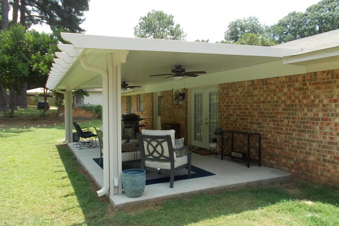White patio cover with ceiling fan shelters a furnished concrete patio beside a brick house