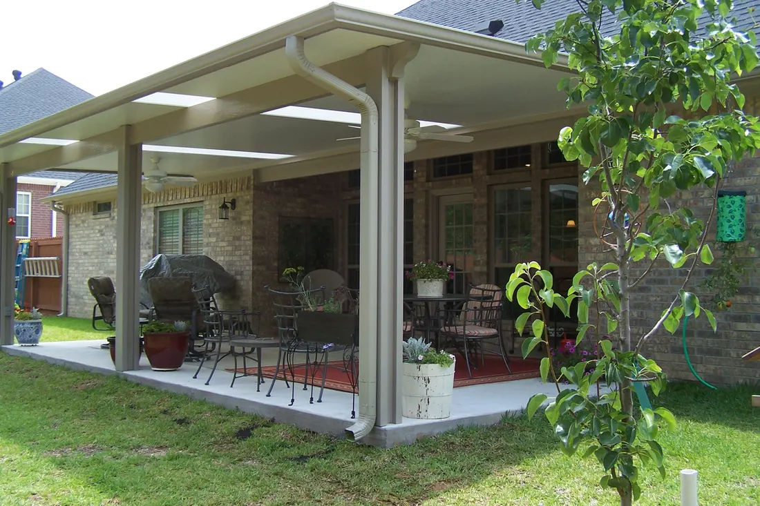 Covered patio with beige roof, white posts, brick walls, outdoor furniture, potted plants, and green lawn